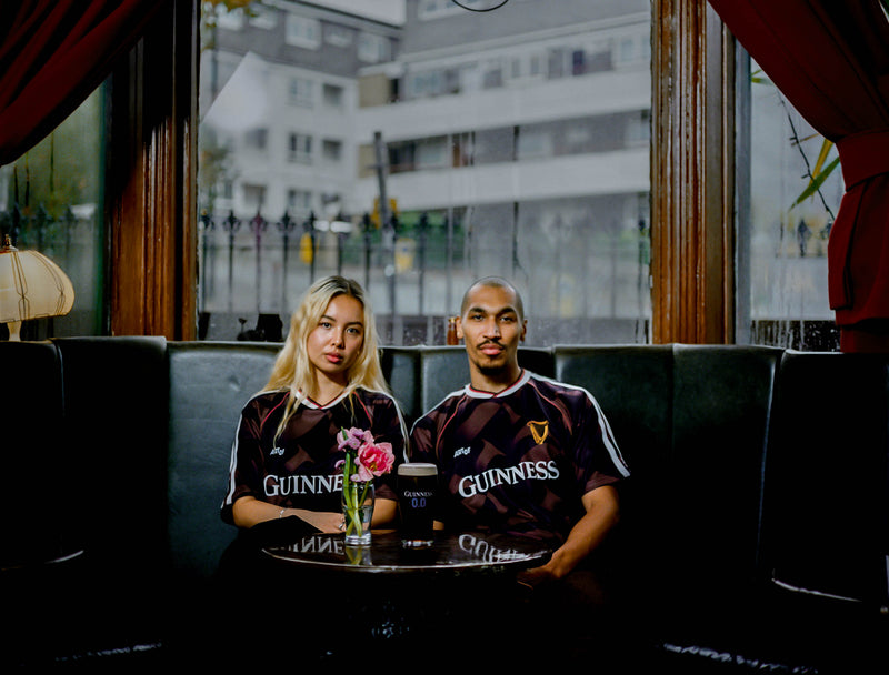 Two people wearing Guinness jerseys sitting on a couch with a table in front of them.