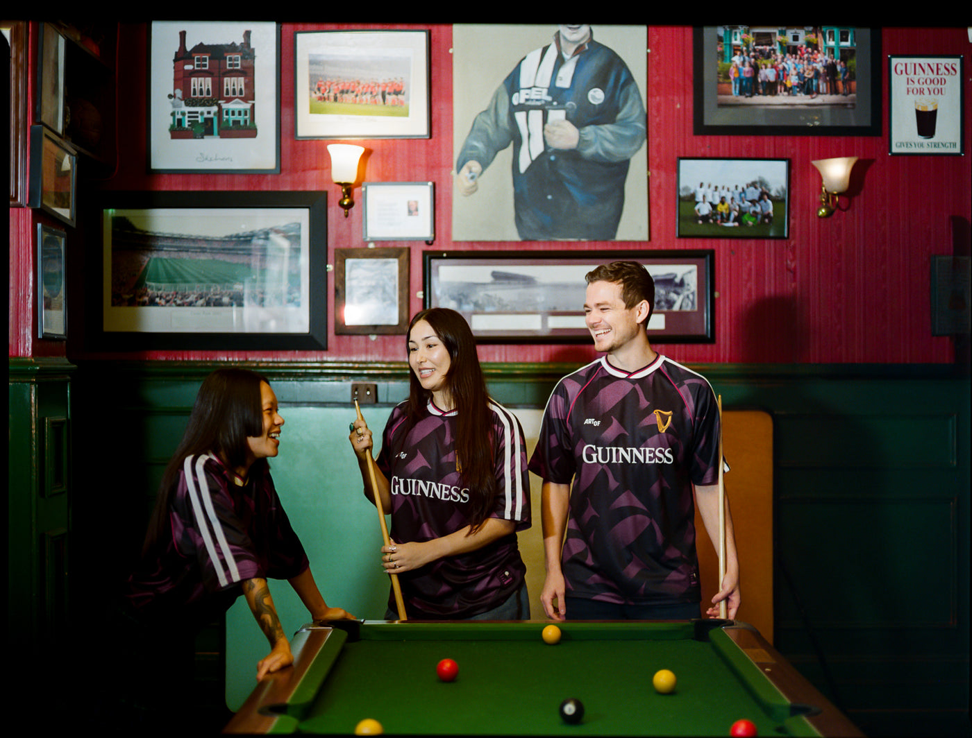 Three people in Guinness-branded jerseys playing pool in a bar with framed pictures on the wall.