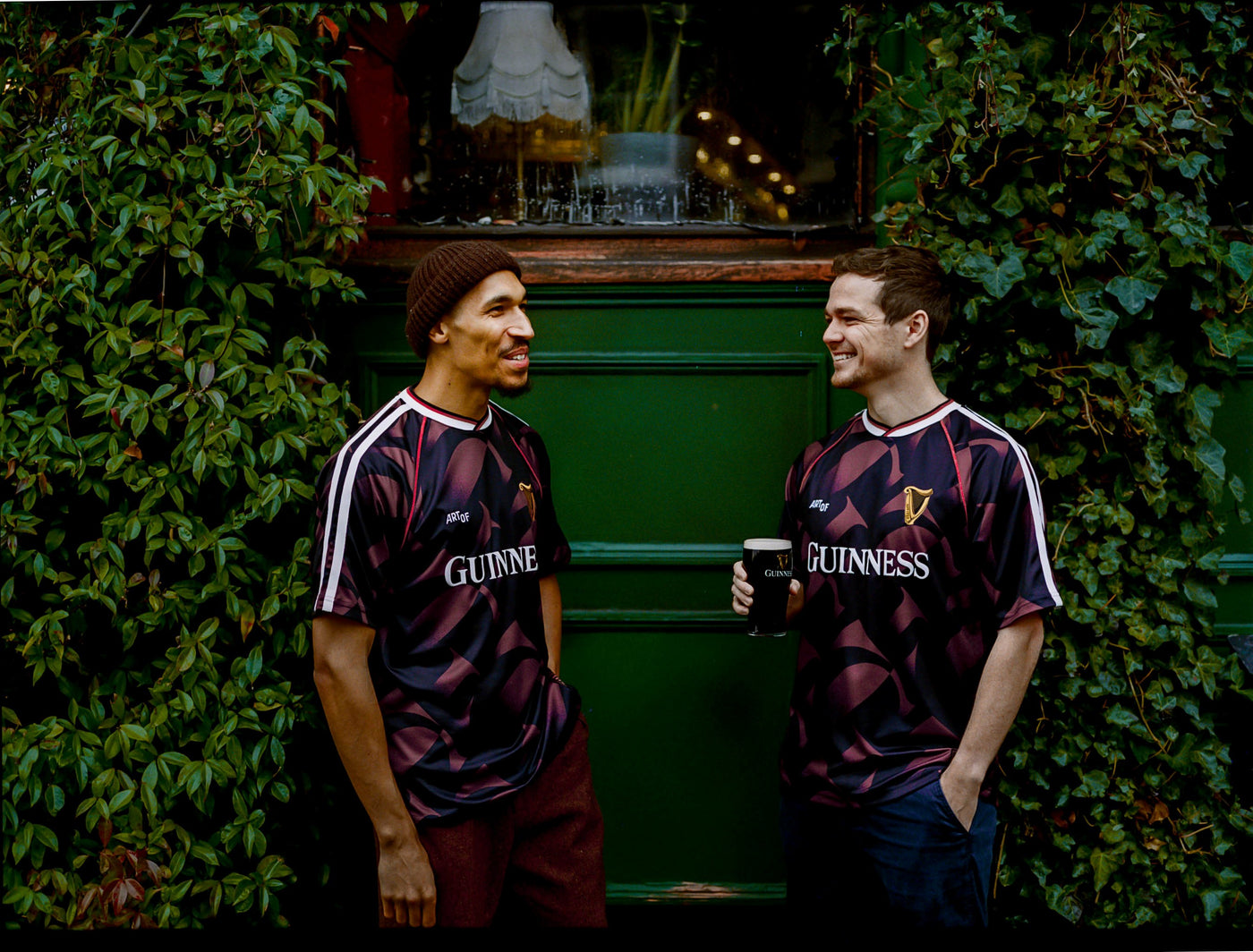 Two men wearing Guinness jerseys standing in front of a green wall with plants.