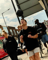Person holding a black medicine ball on a city street with pedestrians and a red bus in the background.